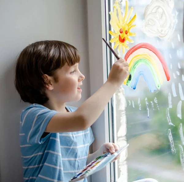 A little boy draws a rainbow on a window during a coronavirus pandemic. Waiting for the end of the COVID-19 epidemic.