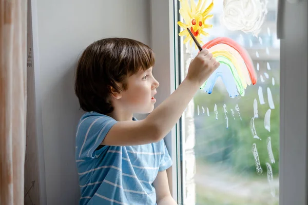 A little boy draws a rainbow on a window during a coronavirus pandemic. Waiting for the end of the COVID-19 epidemic.