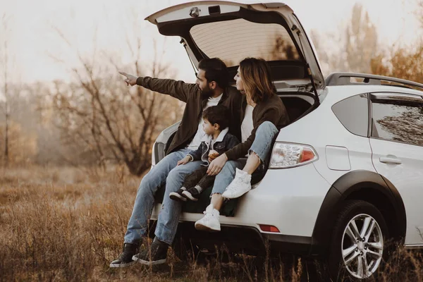 Família feliz sentada no tronco aberto do carro hatchback e apontando o dedo ao ar livre . — Fotografia de Stock