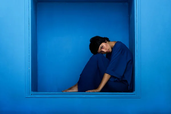 Jovem bela mulher sentada em nicho no fundo azul . — Fotografia de Stock