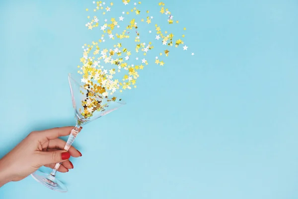 Woman hand holding martini glass with pouring out golden stars confetti on blue background. — Stock Photo, Image