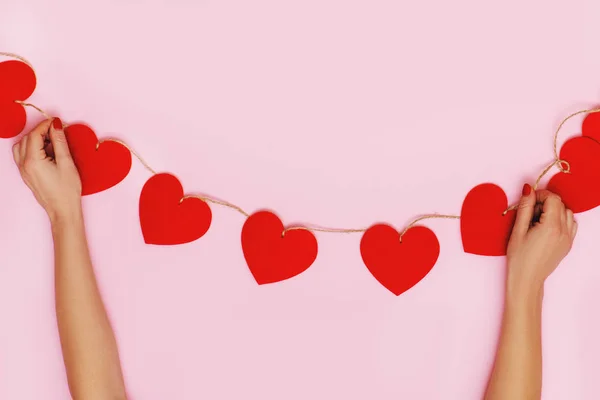 Woman hands holding garland made of hearts on pink background.