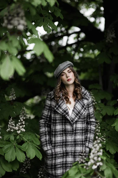 Vacker kvinna i toppad mössa och rutig rock nära blommande kastanjeträd. — Stockfoto