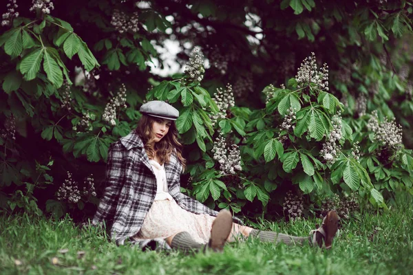 Portrait of beautiful woman in peaked cap, white dress and plaid coat sitting under flowering chestnut tree. — Stock Photo, Image