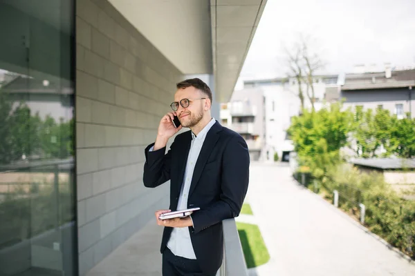 Joven hombre de negocios hipster en gafas hablando de teléfono cerca de la oficina o —  Fotos de Stock