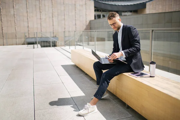 Молодой хипстерский бизнесмен в костюме печатает ноутбук возле офиса outd — стоковое фото