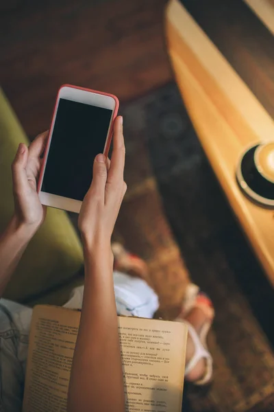 Las manos femeninas sosteniendo el teléfono inteligente y el libro de papel en la cafetería —  Fotos de Stock