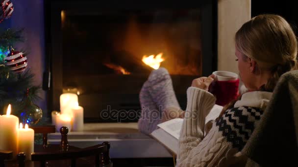 Женщина сидит с чашкой горячего напитка и книгу у камина — стоковое видео