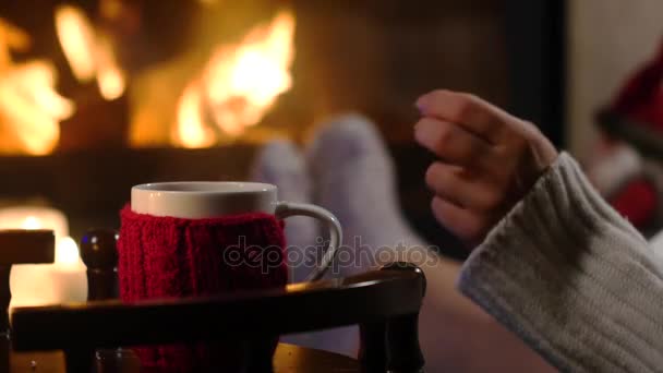 Mujer está sentada con taza de bebida caliente y libro cerca de la chimenea — Vídeos de Stock