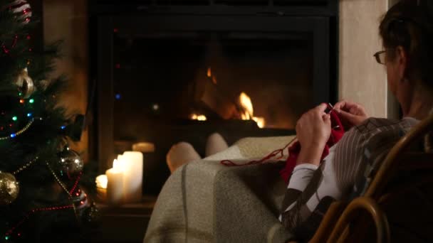 Grandmother knits a sweater sitting in front of a fireplace — Stock Video