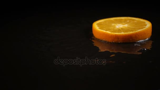 Rodajas de naranja caen sobre la superficie negra cubierta de agua — Vídeos de Stock