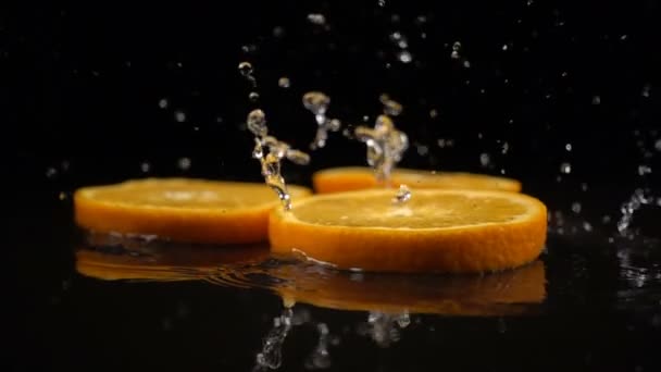 Rodajas de naranja caen sobre la superficie negra cubierta de agua — Vídeo de stock