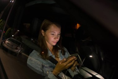 Genç bir kadın gece arabada otururken cep telefonu kullanıyor.