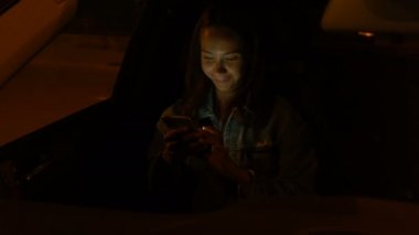 Genç bir kadın gece arabada otururken cep telefonu kullanıyor.
