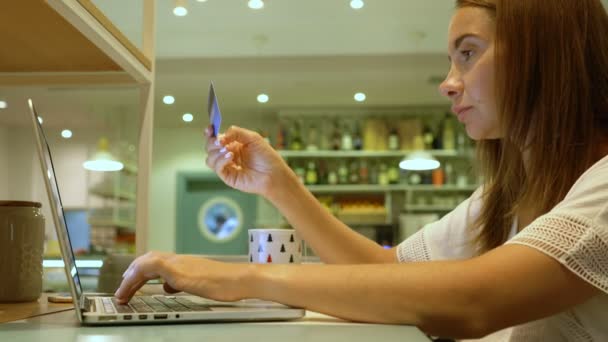 Молодая женщина делает покупки в Интернете, сидя в кафе с банковской картой и ноутбуком — стоковое видео