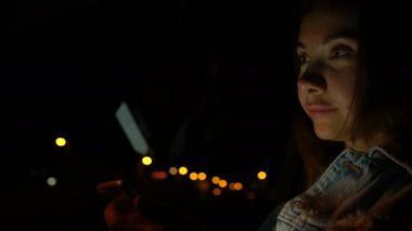 Genç bir kadın gece arabada otururken cep telefonu kullanıyor.