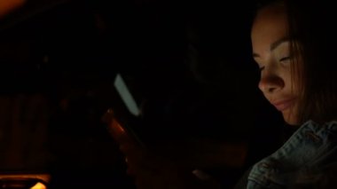 Genç bir kadın gece arabada otururken cep telefonu kullanıyor.