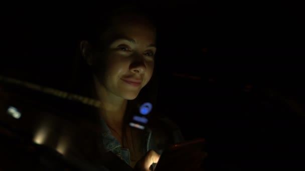 Молодая женщина, сидя ночью в машине, пользуется мобильным телефоном — стоковое видео