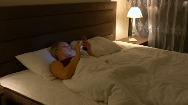 Young woman lies in bed and uses a smartphone in the evening — 비디오