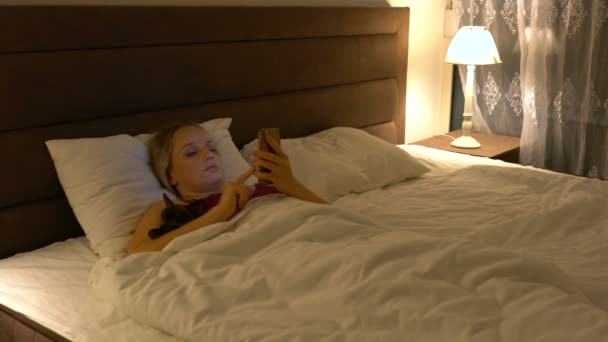 Young woman lies in bed and uses a smartphone in the evening — 비디오