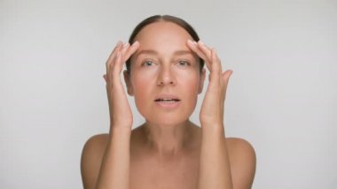 Pürüzsüz, sağlıklı bir cilde sahip genç bir kadının yakın plan güzel portresi, beyaz arka planda parmaklarıyla nazikçe yüzüne dokunur.