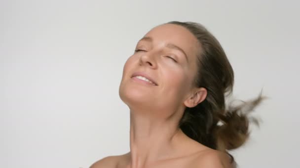 Retrato de belleza de cerca de una mujer joven con una piel suave y saludable, se desata el cabello sobre un fondo blanco y sonríe — Vídeos de Stock