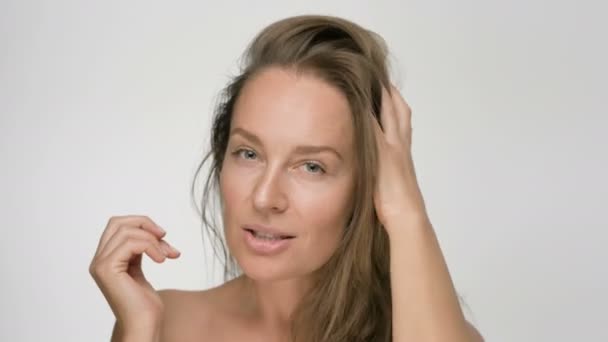 Retrato de belleza de cerca de una mujer joven con una piel suave y saludable, toca suavemente el cabello con las manos y mira juguetonamente a la cámara sobre un fondo blanco — Vídeos de Stock