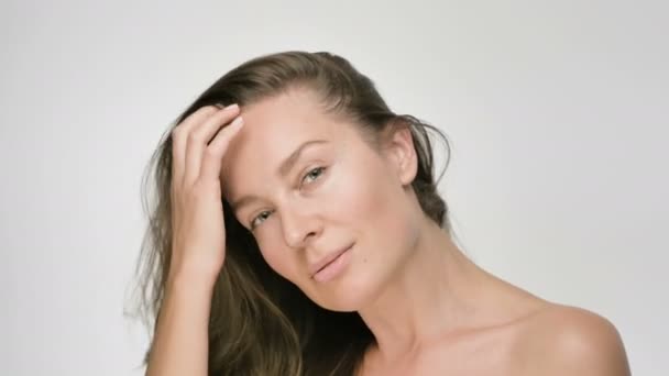 Närbild skönhet porträtt av ung kvinna med slät frisk hud, hon försiktigt vidrör håret med händerna och lekfullt tittar på kameran på vit bakgrund — Stockvideo