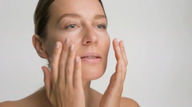 Pürüzsüz, sağlıklı bir cilde sahip genç bir kadının yakın plan güzel portresi, beyaz arka planda parmaklarıyla nazikçe yüzüne dokunur ve gülümser.