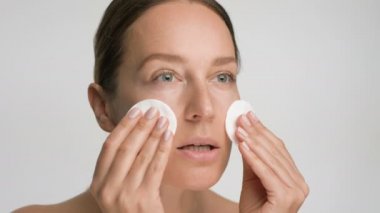 Pürüzsüz, sağlıklı bir cilde sahip genç bir kadının yakın plan güzel portresi, beyaz arka planda pamuk pedlerle yüzünü nazikçe siler.