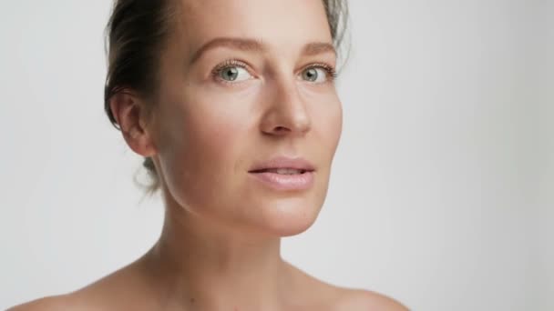 Close-up portret piękna młodej kobiety o gładkiej zdrowej skórze, delikatnie dotyka twarzy palcami na białym tle i uśmiecha — Wideo stockowe