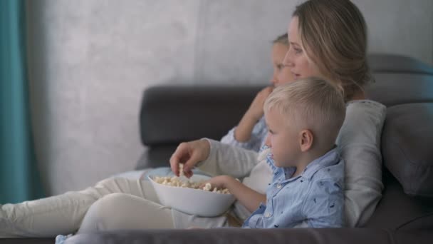 Мама і двоє дітей їдять попкорн і дивляться телевізор, сидячи на дивані — стокове відео
