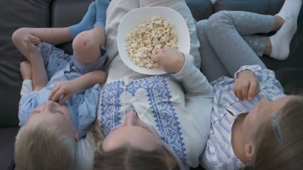 Mama i dwójka dzieci jedzą popcorn i oglądają telewizję siedząc na kanapie — Wideo stockowe