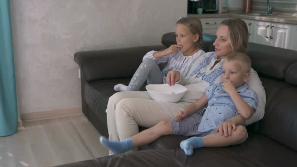 Mamá y dos niños comiendo palomitas de maíz y viendo la televisión mientras están sentados en el sofá — Vídeos de Stock