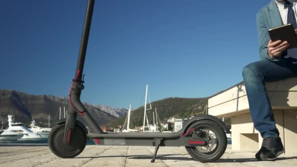 Guapo hombre de negocios sentado al lado de un scooter eléctrico en un banco sonriendo y trabajando usando una tableta — Vídeo de stock