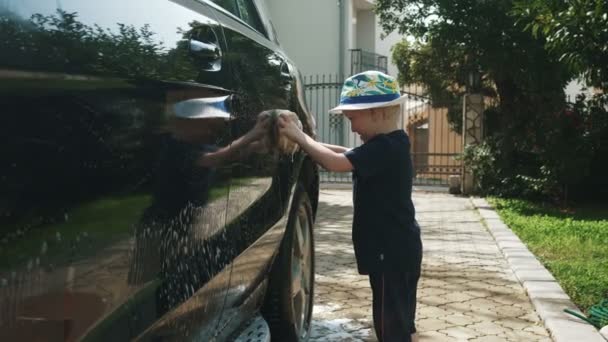 Kleine jongen in een hoed wast een auto met zeep en een spons — Stockvideo