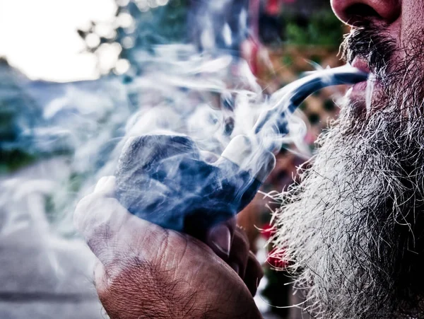 happy old man smokes the tobacco in his pipe with pleasure among sweet swirls and clouds of smoke