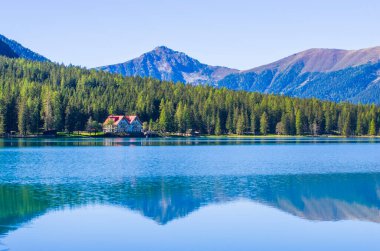 Saf mavi suları olan dağ gölü, tarım kulübesi ve açık gökyüzünün altındaki yeşil orman.