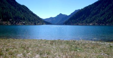 Mavi gökyüzünün altında parlak mavi dağ gölü ve etrafındaki yeşil orman ve granit taşlar ve tepesinde kar