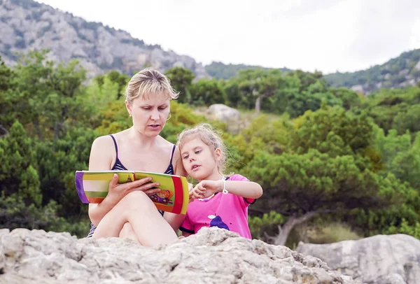 De dochter luistert naar haar moeder die haar in de zomer een boek voorleest in de natuur. — Stockfoto