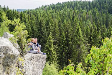 Bir erkek ve bir kadın, doğadaki bir dağın kenarında oturup birbirlerine sarılırlar..