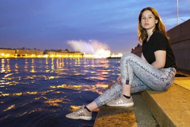 Kız, Neva nehrinin setindeki akşam kentin arka planındaki granit basamaklarda oturuyor. Saint-Petersburg.