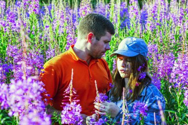 Genç bir kız ve bir adam uzun otların arasında çiçeklerle sarılıyorlar.