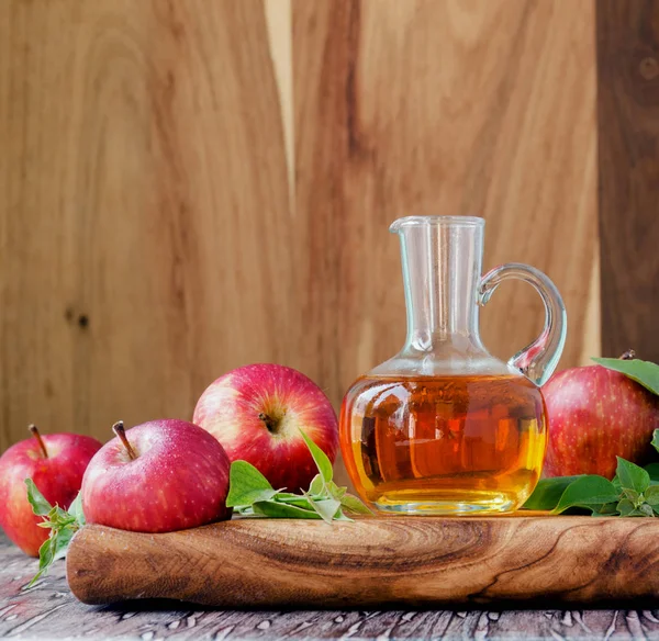 Glaskanna med äppelcidervinäger och mogna röda äpplen på träbord, selektivt fokus — Stockfoto