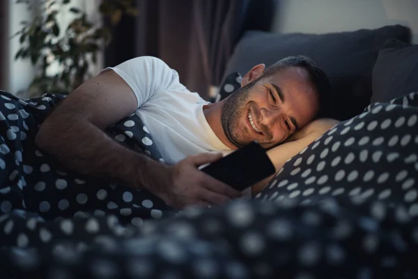 Joven adicto hombre despierto en la noche en la cama usando teléfono inteligente —  Fotos de Stock