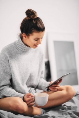 Kız sabah yatağın üstüne oturan ve haber okumak için tablet kullanma 