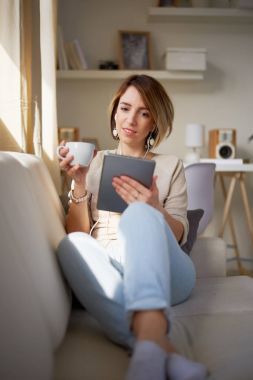 Fincan çay ve dijital tablet ile kanepede oturan genç güzel kadın