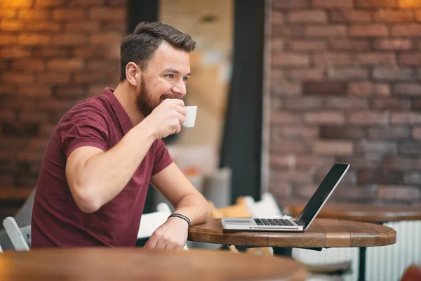 Hombre bebiendo café y usando el ordenador portátil mientras está sentado en la cafetería . — Foto de Stock
