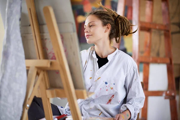 Artistic girl sitting in studio and paint on easel. — Stock Photo, Image