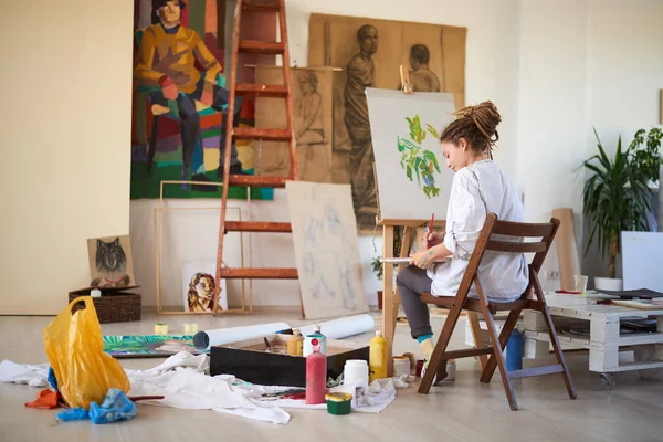 Menina artística sentado em estúdio e pintura no cavalete . — Fotografia de Stock